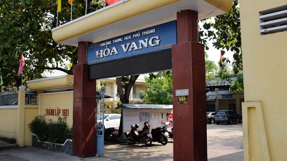 Entrance gate of a school labeled 'Hòa Vang' with large pillars and a sign displaying the school's name. Trees provide shade over the entrance, and there are motorcycles parked in the area. A yellow wall runs alongside, featuring some text. In the background, parts of buildings and a car are visible, indicating an urban setting.