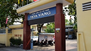 Entrance gate of a school labeled 'H&ograve;a Vang' with large pillars and a sign displaying the school's name. Trees provide shade over the entrance, and there are motorcycles parked in the area. A yellow wall runs alongside, featuring some text. In the background, parts of buildings and a car are visible, indicating an urban setting.