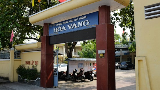 Entrance gate of a school labeled 'H&ograve;a Vang' with large pillars and a sign displaying the school's name. Trees provide shade over the entrance, and there are motorcycles parked in the area. A yellow wall runs alongside, featuring some text. In the background, parts of buildings and a car are visible, indicating an urban setting.