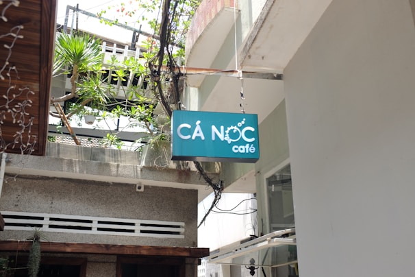 A teal sign with white lettering displaying the name 'Cá Nóc café' hangs outside a building, surrounded by potted plants and wires, with a partially visible balcony above.