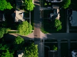 Aerial view of a residential area with several houses aligned along streets. Lush green trees surround the homes, casting shadows over the roads. The layout includes driveways and walkways leading up to each house, with patches of grass and gardens contributing to the verdant landscape. A single vehicle is parked on a driveway.