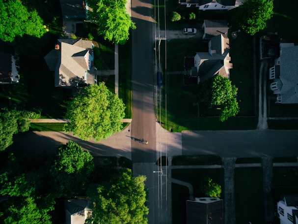 Aerial view of a residential area with several houses aligned along streets. Lush green trees surround the homes, casting shadows over the roads. The layout includes driveways and walkways leading up to each house, with patches of grass and gardens contributing to the verdant landscape. A single vehicle is parked on a driveway.