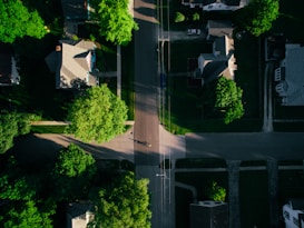 Aerial view of a residential area with several houses aligned along streets. Lush green trees surround the homes, casting shadows over the roads. The layout includes driveways and walkways leading up to each house, with patches of grass and gardens contributing to the verdant landscape. A single vehicle is parked on a driveway.