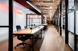 A modern office space featuring an open-plan layout with long wooden desks and black office chairs. The interior includes glass walls with black frames and overhead industrial-style lighting. A red neon sign provides ambient lighting, and exposed ceiling ductwork adds to the industrial aesthetic.