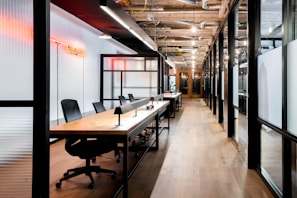 A modern office space featuring an open-plan layout with long wooden desks and black office chairs. The interior includes glass walls with black frames and overhead industrial-style lighting. A red neon sign provides ambient lighting, and exposed ceiling ductwork adds to the industrial aesthetic.