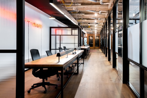 A modern office space featuring an open-plan layout with long wooden desks and black office chairs. The interior includes glass walls with black frames and overhead industrial-style lighting. A red neon sign provides ambient lighting, and exposed ceiling ductwork adds to the industrial aesthetic.