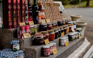 A market stall displays an assortment of bottled goods including wines, various jars of preserves, sauces, and vegetables. Price tags are placed in front of each item. The items are organized on a multi-tiered wooden display. In the background, a white van and some greenery can be seen.