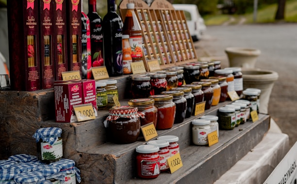 A market stall displays an assortment of bottled goods including wines, various jars of preserves, sauces, and vegetables. Price tags are placed in front of each item. The items are organized on a multi-tiered wooden display. In the background, a white van and some greenery can be seen.