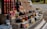 A market stall displays an assortment of bottled goods including wines, various jars of preserves, sauces, and vegetables. Price tags are placed in front of each item. The items are organized on a multi-tiered wooden display. In the background, a white van and some greenery can be seen.