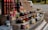 A market stall displays an assortment of bottled goods including wines, various jars of preserves, sauces, and vegetables. Price tags are placed in front of each item. The items are organized on a multi-tiered wooden display. In the background, a white van and some greenery can be seen.