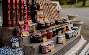 A market stall displays an assortment of bottled goods including wines, various jars of preserves, sauces, and vegetables. Price tags are placed in front of each item. The items are organized on a multi-tiered wooden display. In the background, a white van and some greenery can be seen.