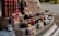 A market stall displays an assortment of bottled goods including wines, various jars of preserves, sauces, and vegetables. Price tags are placed in front of each item. The items are organized on a multi-tiered wooden display. In the background, a white van and some greenery can be seen.