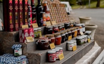 A market stall displays an assortment of bottled goods including wines, various jars of preserves, sauces, and vegetables. Price tags are placed in front of each item. The items are organized on a multi-tiered wooden display. In the background, a white van and some greenery can be seen.