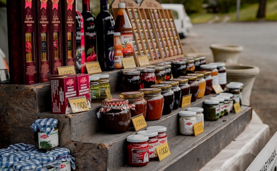 A market stall displays an assortment of bottled goods including wines, various jars of preserves, sauces, and vegetables. Price tags are placed in front of each item. The items are organized on a multi-tiered wooden display. In the background, a white van and some greenery can be seen.