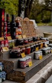 A rustic outdoor market stand displays a variety of bottled products, including jams, sauces, and wines. Jars and bottles are arranged neatly on wooden shelves with handwritten price tags in front. The setting suggests a countryside environment with green foliage in the background, adding a natural, serene atmosphere.
