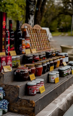 A rustic outdoor market stand displays a variety of bottled products, including jams, sauces, and wines. Jars and bottles are arranged neatly on wooden shelves with handwritten price tags in front. The setting suggests a countryside environment with green foliage in the background, adding a natural, serene atmosphere.