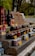 A rustic outdoor market stand displays a variety of bottled products, including jams, sauces, and wines. Jars and bottles are arranged neatly on wooden shelves with handwritten price tags in front. The setting suggests a countryside environment with green foliage in the background, adding a natural, serene atmosphere.