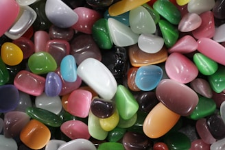 A collection of polished, multicolored stones of varying shapes and sizes, with smooth surfaces reflecting light. The stones feature a range of vibrant colors including greens, pinks, blues, oranges, and whites, arranged in a random close-up display.