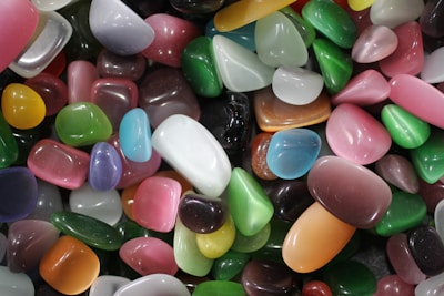 A collection of polished, multicolored stones of varying shapes and sizes, with smooth surfaces reflecting light. The stones feature a range of vibrant colors including greens, pinks, blues, oranges, and whites, arranged in a random close-up display.