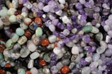 A colorful assortment of polished gemstones, featuring a variety of shapes and sizes. The stones display a spectrum of colors including purples, greens, reds, and blues, reflecting light with their smooth, shiny surfaces.