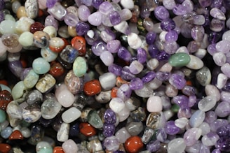 A colorful assortment of polished gemstones, featuring a variety of shapes and sizes. The stones display a spectrum of colors including purples, greens, reds, and blues, reflecting light with their smooth, shiny surfaces.