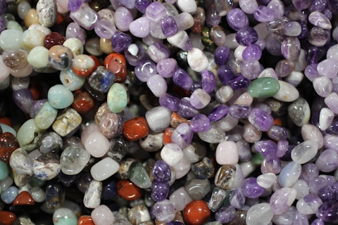 A colorful assortment of polished gemstones, featuring a variety of shapes and sizes. The stones display a spectrum of colors including purples, greens, reds, and blues, reflecting light with their smooth, shiny surfaces.