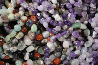 A colorful assortment of polished gemstones, featuring a variety of shapes and sizes. The stones display a spectrum of colors including purples, greens, reds, and blues, reflecting light with their smooth, shiny surfaces.