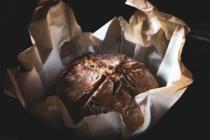 A rustic loaf of bread is nestled in crinkled parchment paper. The bread has a brown, textured crust with visible cracks, suggesting a crunchy exterior.