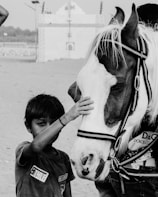 A young boy gently pets a horse on the nose. The setting appears to be outdoors, possibly in a desert or sandy area, with a building visible in the background. The image is in black and white, emphasizing textures and contrasts.