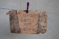 A handmade sign with the message 'Creativity is better left unconfined' is attached to a weathered, textured background using a purple clothespin on a blue and white rope. The sign blends an artistic mix of handwritten styles in a combination of black and some colorful accents.