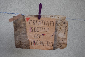A handmade sign with the message 'Creativity is better left unconfined' is attached to a weathered, textured background using a purple clothespin on a blue and white rope. The sign blends an artistic mix of handwritten styles in a combination of black and some colorful accents.