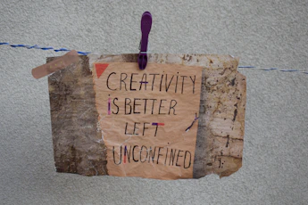 A handmade sign with the message 'Creativity is better left unconfined' is attached to a weathered, textured background using a purple clothespin on a blue and white rope. The sign blends an artistic mix of handwritten styles in a combination of black and some colorful accents.