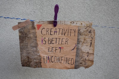 A handmade sign with the message 'Creativity is better left unconfined' is attached to a weathered, textured background using a purple clothespin on a blue and white rope. The sign blends an artistic mix of handwritten styles in a combination of black and some colorful accents.