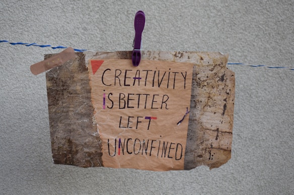 A handmade sign with the message 'Creativity is better left unconfined' is attached to a weathered, textured background using a purple clothespin on a blue and white rope. The sign blends an artistic mix of handwritten styles in a combination of black and some colorful accents.