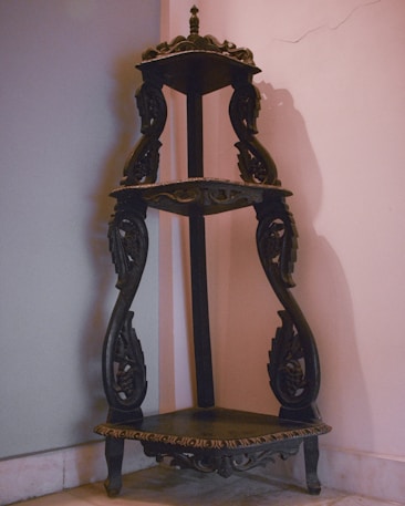 An ornate, dark wooden corner shelf featuring intricate carvings and a three-tiered design. The structure is placed against a wall with one section showing a pale pink hue and the other a light gray shade. The craftsmanship displays detailed scrollwork and delicate patterns, adding an antique and elegant appearance.