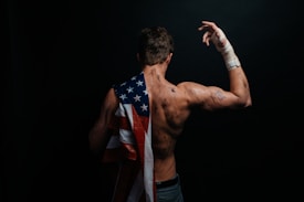 A muscular person with a rugged, athletic appearance is seen from behind, draped in a US flag. Their right arm is bent, with a taped wrist and hand, suggesting they've been engaged in intense physical activity. The skin is marked with dirt or bruises, conveying a sense of struggle or endurance.