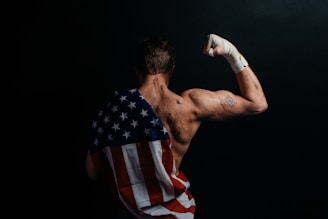 A muscular person stands with their back to the camera, flexing their arm. The skin appears sweaty or marked, and a bandaged fist is raised. Draped across the back is a cloth resembling the American flag, emphasizing strength and patriotism. The background is dark, enhancing the focus on the person.