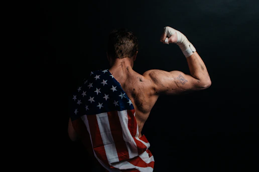 A muscular person stands with their back to the camera, flexing their arm. The skin appears sweaty or marked, and a bandaged fist is raised. Draped across the back is a cloth resembling the American flag, emphasizing strength and patriotism. The background is dark, enhancing the focus on the person.