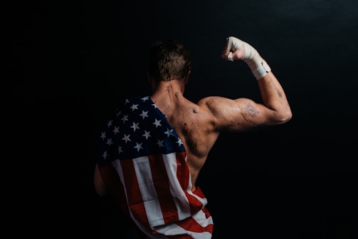 A muscular person stands with their back to the camera, flexing their arm. The skin appears sweaty or marked, and a bandaged fist is raised. Draped across the back is a cloth resembling the American flag, emphasizing strength and patriotism. The background is dark, enhancing the focus on the person.