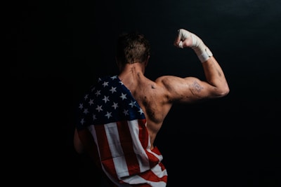 A muscular person stands with their back to the camera, flexing their arm. The skin appears sweaty or marked, and a bandaged fist is raised. Draped across the back is a cloth resembling the American flag, emphasizing strength and patriotism. The background is dark, enhancing the focus on the person.