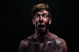 A person with a shocked or terrified expression, covered in dark paint or makeup, with the background in shadow. Their eyes are wide open and mouth agape, conveying a sense of horror or surprise.
