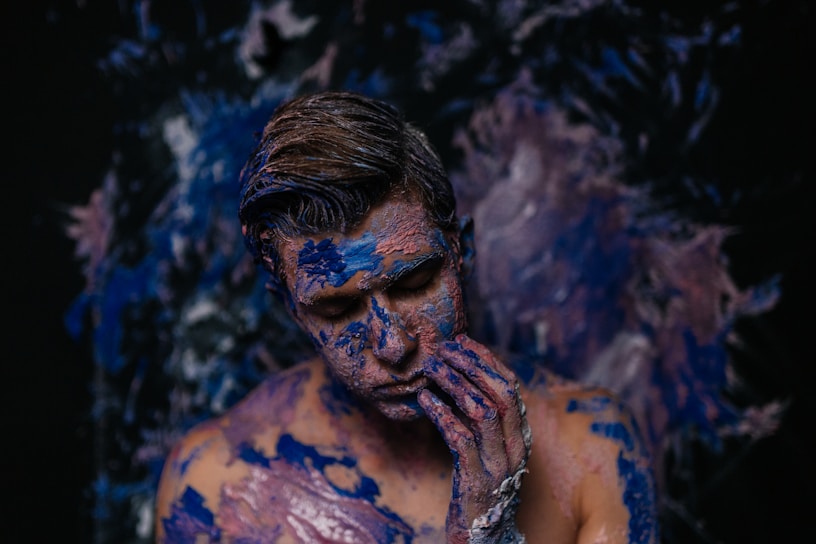 A person is covered in blue and purple paint against a dark, abstract background. The individual's eyes are closed, and their fingers gently touch their face, contributing to an artistic and introspective atmosphere.