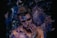A person is covered in blue and purple paint against a dark, abstract background. The individual's eyes are closed, and their fingers gently touch their face, contributing to an artistic and introspective atmosphere.