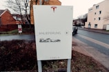 A sign with the Tesla logo on top is placed next to a road. The sign contains German text that reads 'Anlieferung', indicating delivery, with an illustration of a truck carrying a vehicle. Behind the sign, there are residential buildings, trees without leaves, and a wet road suggesting recent rain.