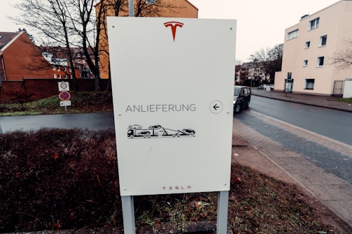 A sign with the Tesla logo on top is placed next to a road. The sign contains German text that reads 'Anlieferung', indicating delivery, with an illustration of a truck carrying a vehicle. Behind the sign, there are residential buildings, trees without leaves, and a wet road suggesting recent rain.