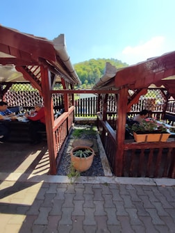 Wooden gazebos with lattice walls house several people who are seated and possibly dining. There is a clay pot with plants in the center walkway between the gazebos. In the background, a wooden fence and a lush, green hill under a bright blue sky can be seen.