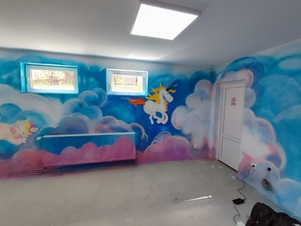 A room with walls painted in a fantasy theme featuring blue and pink clouds. On the wall, there are artistic depictions of unicorns, one flying with a rainbow-colored mane. The room has two small windows with natural light filtering through and a heater painted in a similar theme.