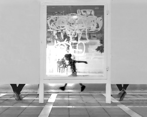 A black and white photograph captures the silhouette of a person running, visible through a glass panel with graffiti. Below the panel, legs of three other people are visible, suggesting they are standing or sitting on the other side. The urban setting with a tiled pavement adds to the dynamic composition.