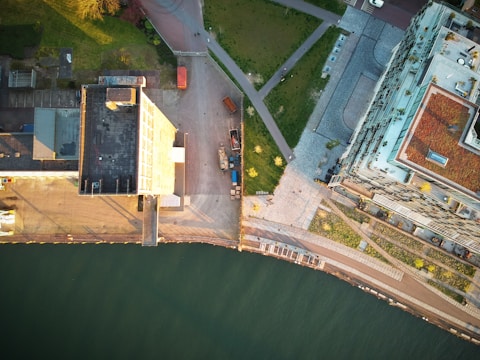 An aerial view of an urban area featuring tall buildings, a body of water, green spaces, and pathways. There are shadows cast by the structures, creating a play of light and shade across the scene, with some small vehicles or containers visible.