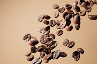 A collection of coffee beans suspended in mid-air against a light brown background. The beans are rich, glossy, and variably textured, showcasing different shades and highlights.